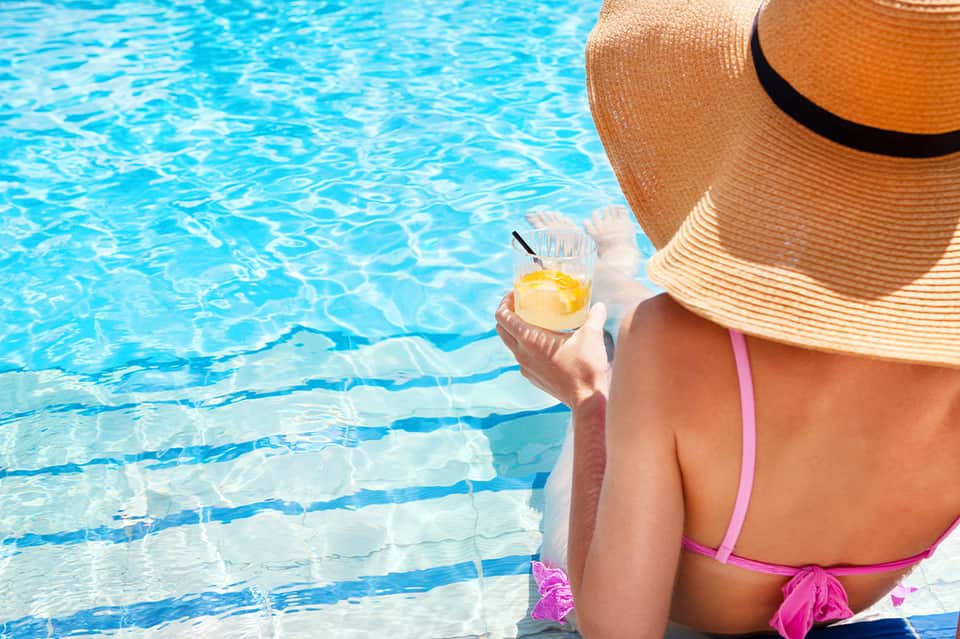 Woman poolside wearing pink bikini and straw hat, holding glass of yellow beverage