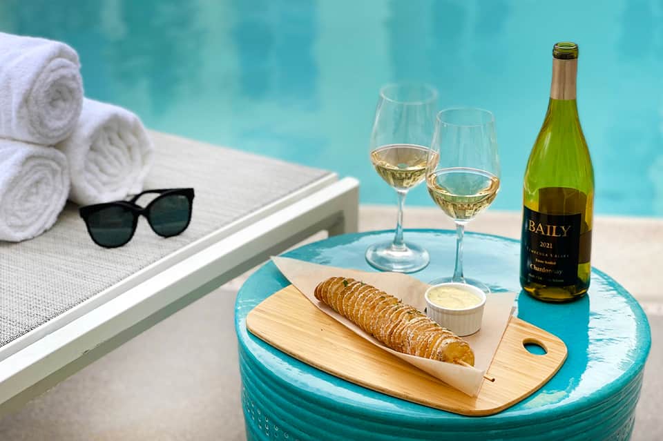 Poolside table with white wine bottle, wine glasses, bread, and sunglasses beside turquoise water
