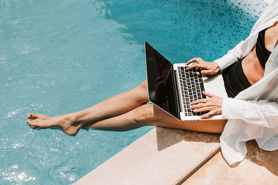Person working on laptop by turquoise swimming pool edge with feet in water