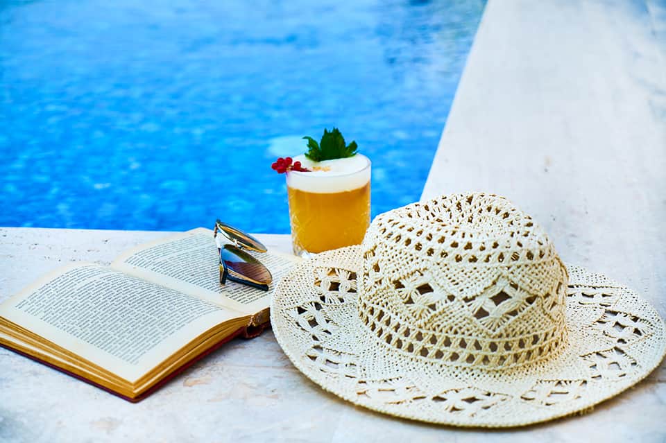Poolside scene with straw hat, sunglasses, open book, and tropical cocktail by turquoise water