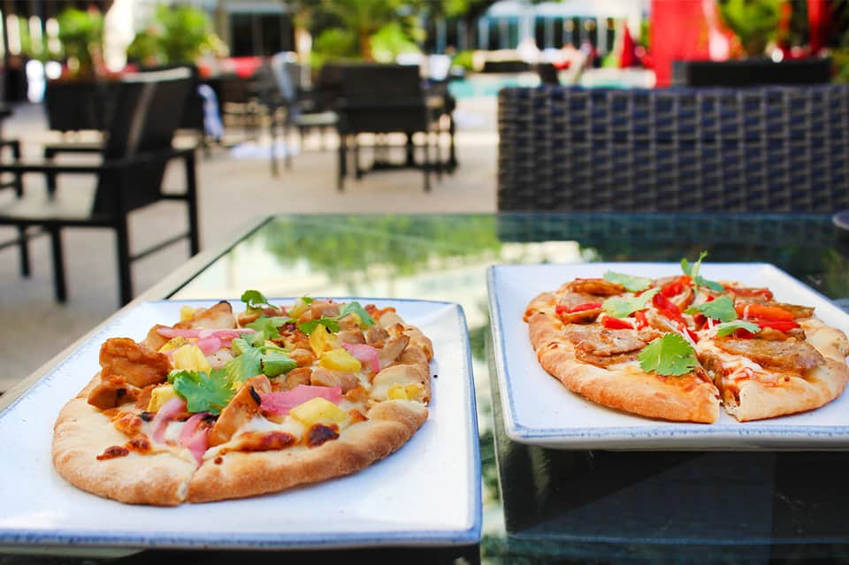 Two gourmet pizzas on white plates served at poolside dining area with blurred seating in background