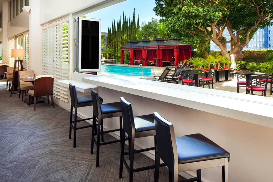 Modern hotel bar overlooking resort pool with red cabanas and lush landscaping