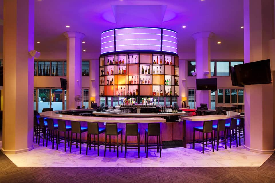 Upscale nightclub lounge with purple ambient lighting, circular bottle display bar, and modern seating