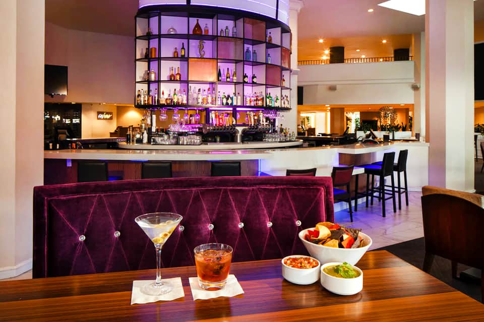 Modern hotel lounge with purple velvet seating, illuminated bottle display, and cocktails with appetizers