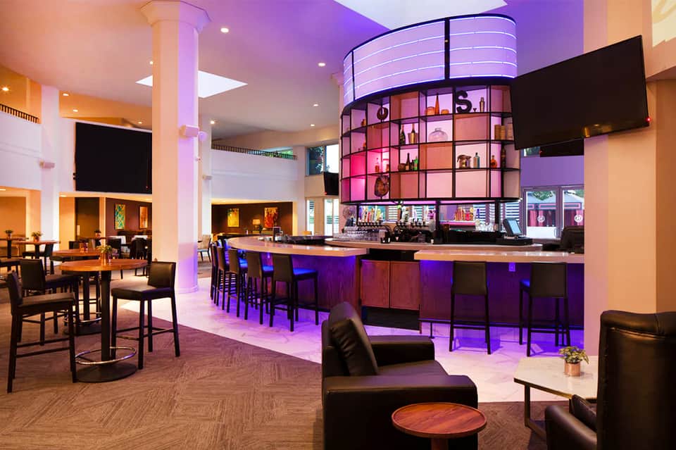 Modern hotel bar lounge with purple ambient lighting, circular shelving, black bar seating, and dining area