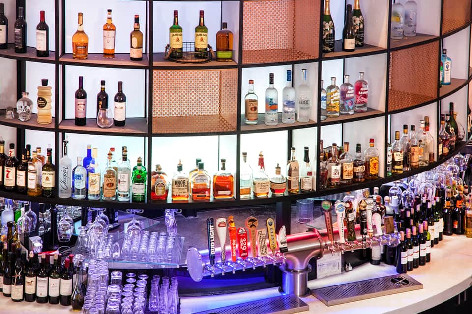 Illuminated bar display with curved shelving, hundreds of liquor bottles, blue ambient lighting, and orange decorative panels