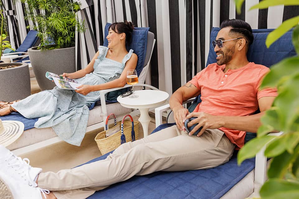 Couple relaxing on lounge chairs under striped cabana with beverages and magazine