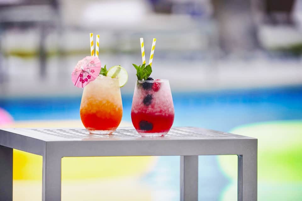 Two colorful tropical cocktails with striped straws and garnishes displayed by a resort pool