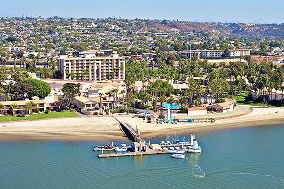 Beachfront resort with multi-story hotel building, palm trees, and research vessel moored offshore