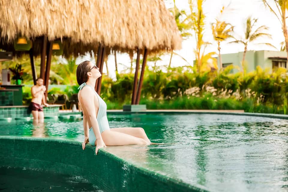 Woman in white swimsuit sitting at edge of tropical infinity pool with thatched-roof structures and palm trees