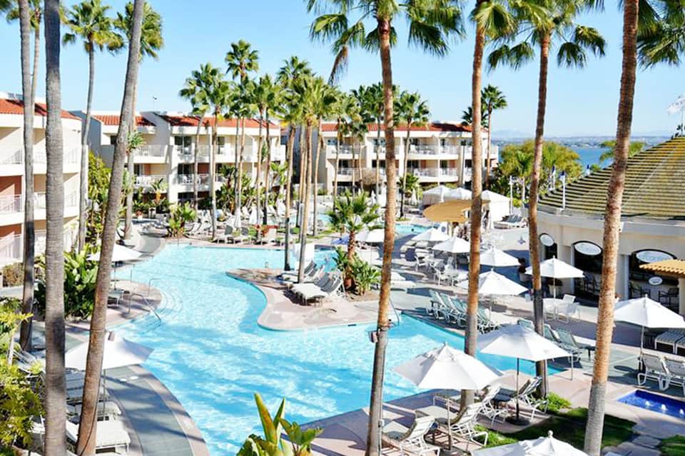 Resort courtyard with curved blue swimming pool, palm trees, and multi-story white buildings under clear sky