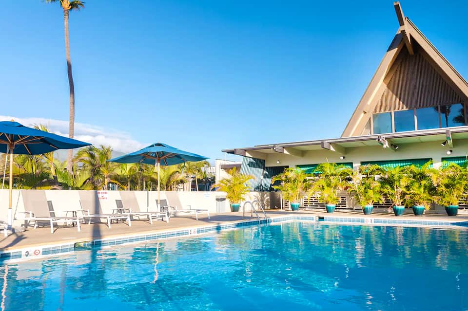 Resort pool with blue umbrellas, white loungers, palm trees, and distinctive A-frame building architecture