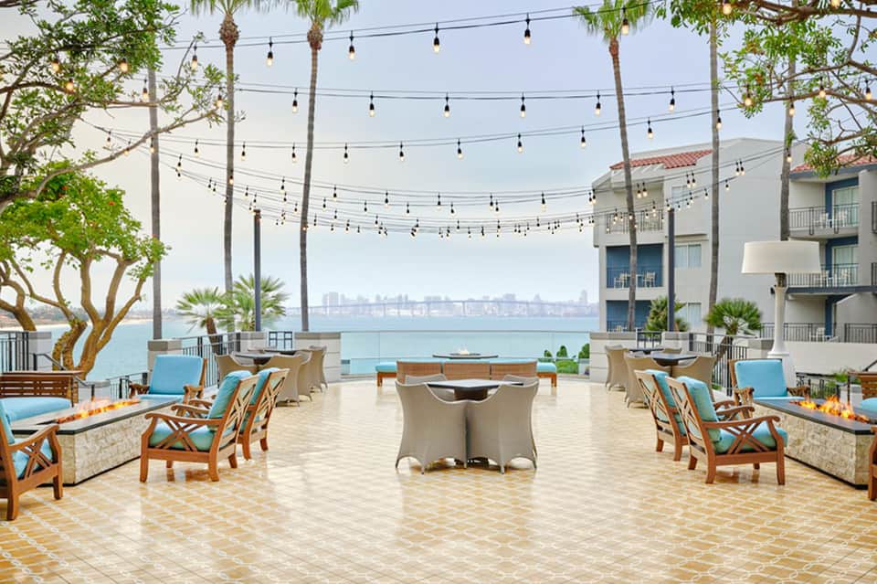 Beachfront terrace with fire pits, wooden lounge chairs, string lights, and ocean view of cityscape