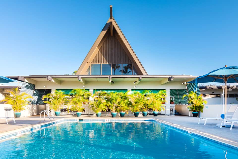 Resort swimming pool with A-frame clubhouse, palm trees, blue umbrellas, and clear skies