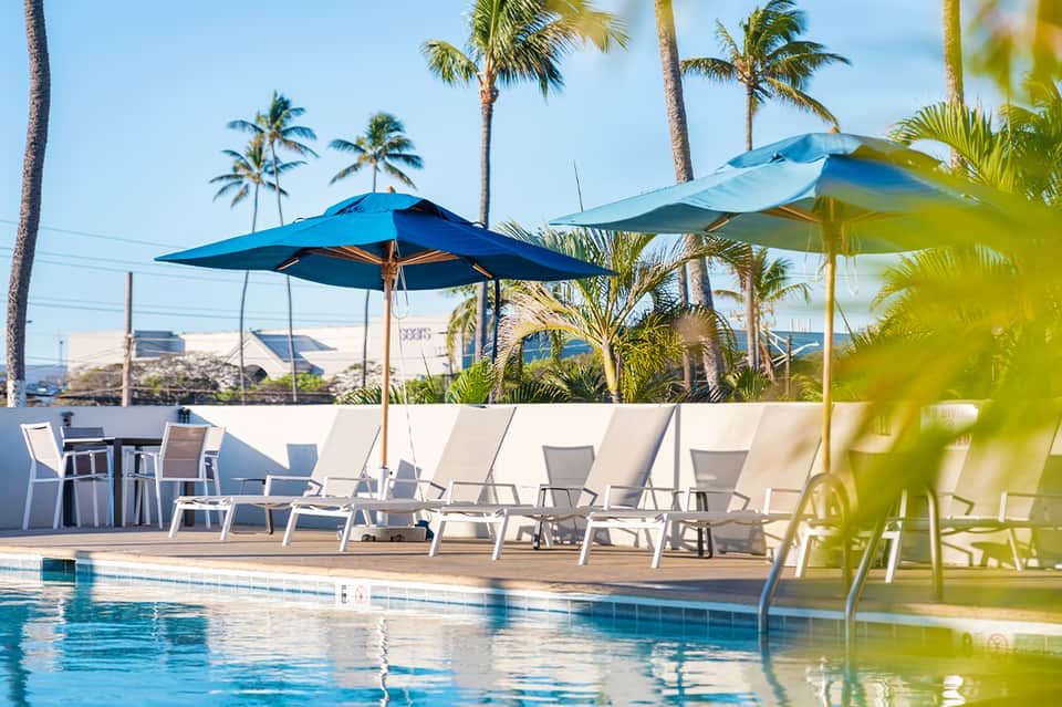 Resort pool deck with blue umbrellas, lounging chairs, and palm trees under clear sky