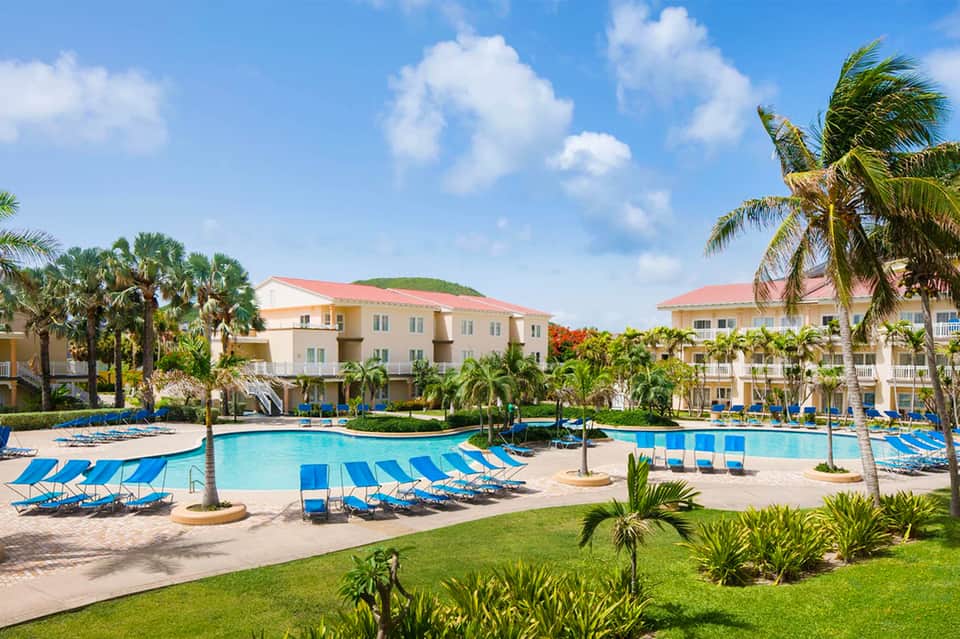 Large resort pool with blue lounge chairs, palm trees, and colorful residential buildings