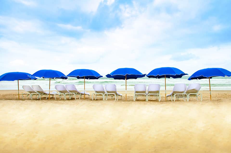 Beach setup with rows of white lounge chairs under blue umbrellas facing the ocean