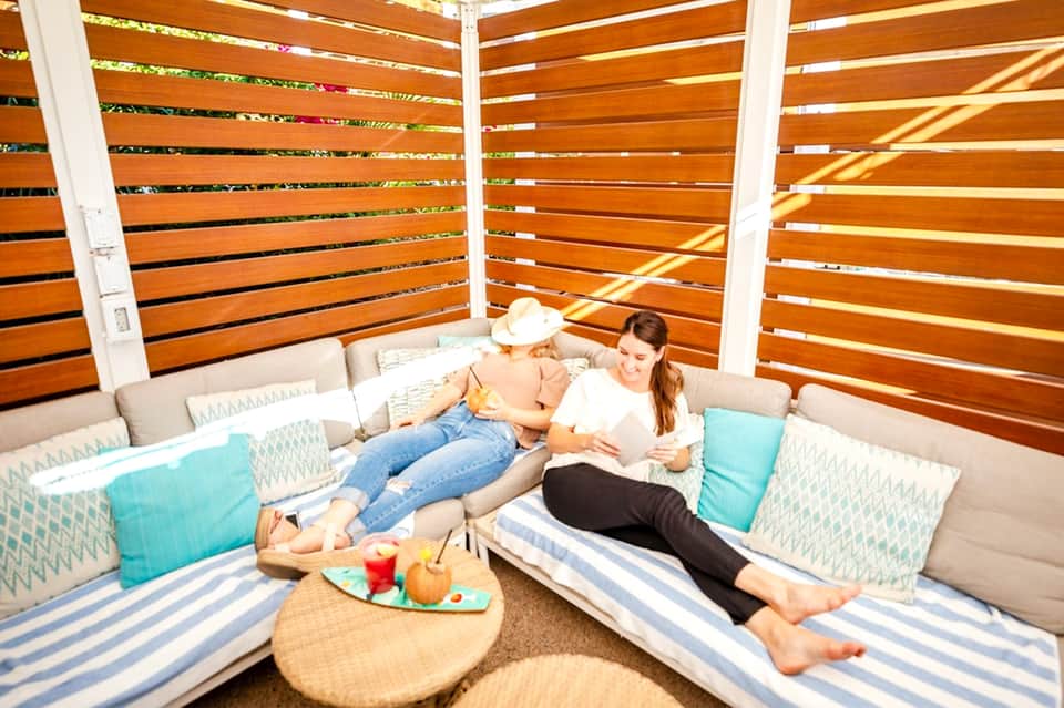 Two women lounging in wooden cabana with shaded seating and fresh fruit