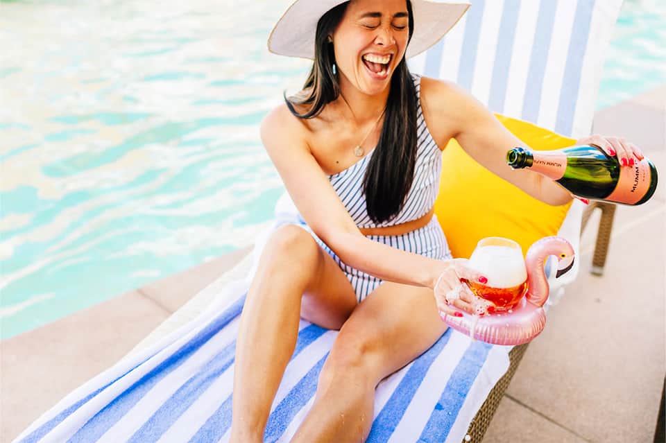 Woman in striped swimsuit pouring champagne by pool with flamingo float