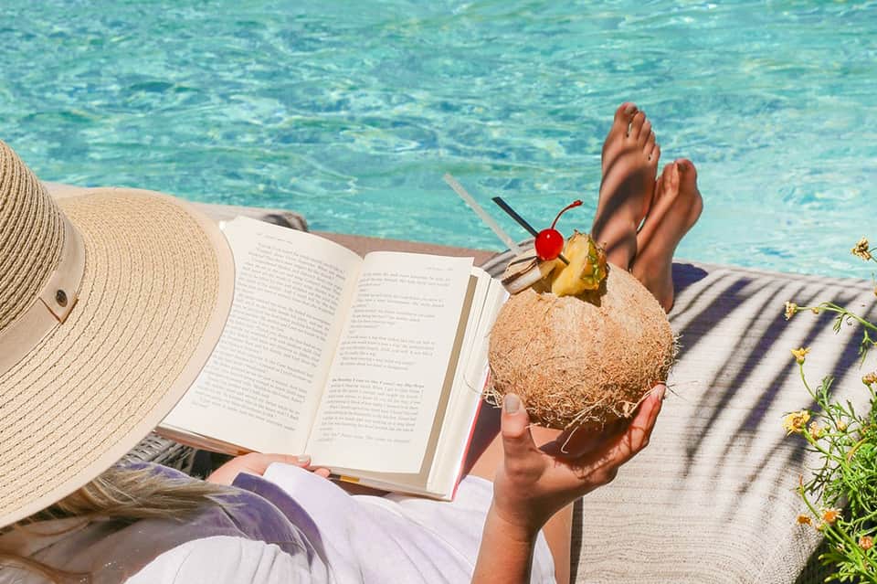 Woman relaxing poolside reading a book while holding a coconut drink