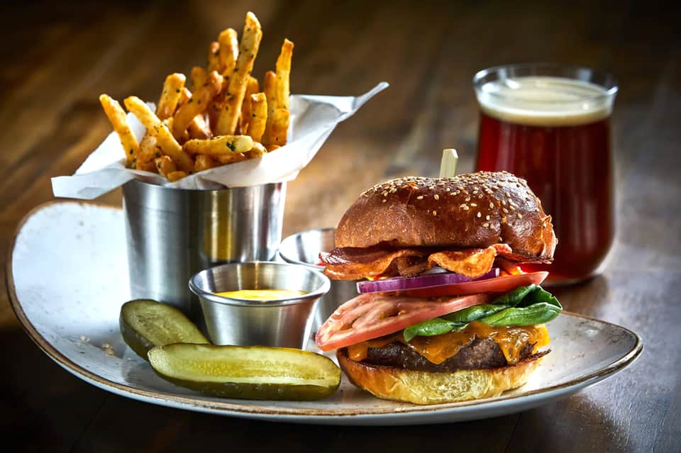 Gourmet burger meal with fries, pickle, dipping sauce, and beer on wooden table