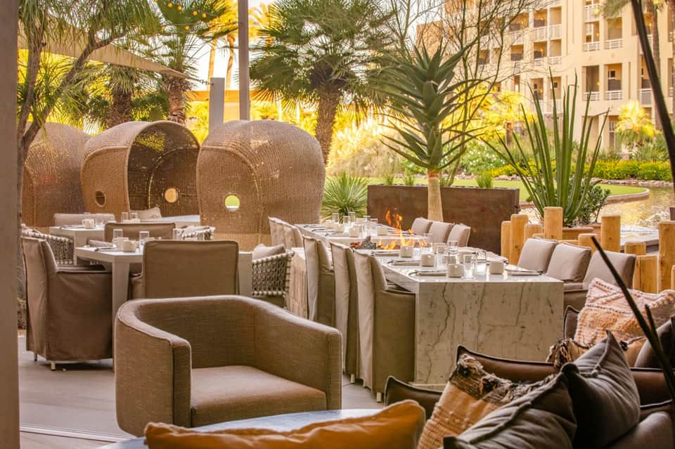 Outdoor dining area with woven dome-shaped lounge chairs, fire pits, palm trees, and modern hotel buildings