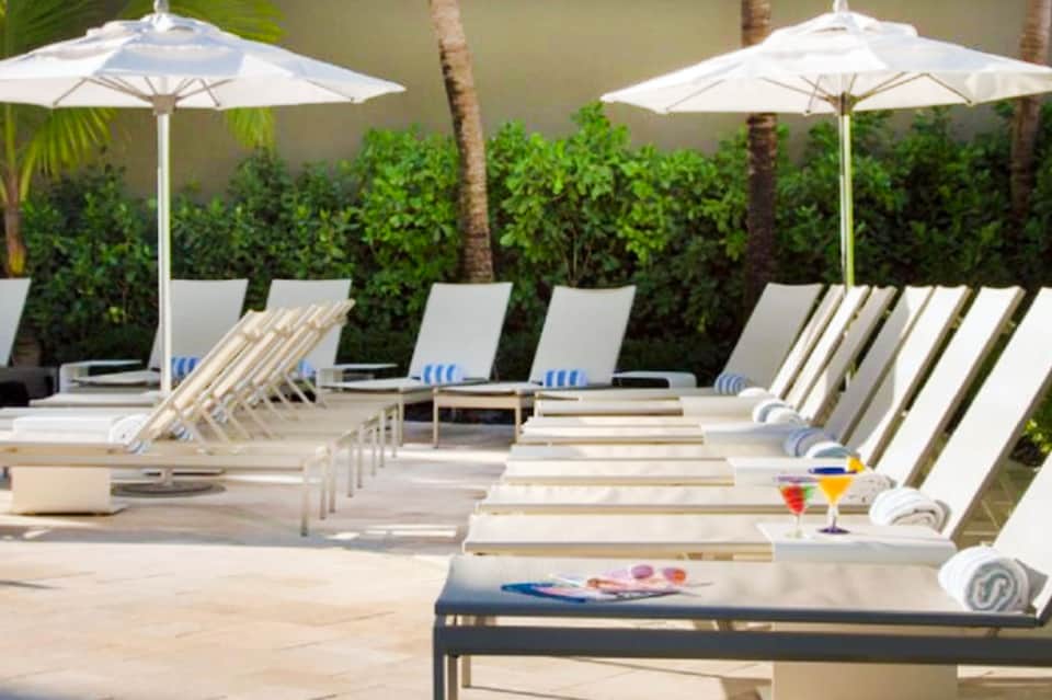 Outdoor pool deck with rows of white lounge chairs, umbrellas, and tropical hedge privacy screening