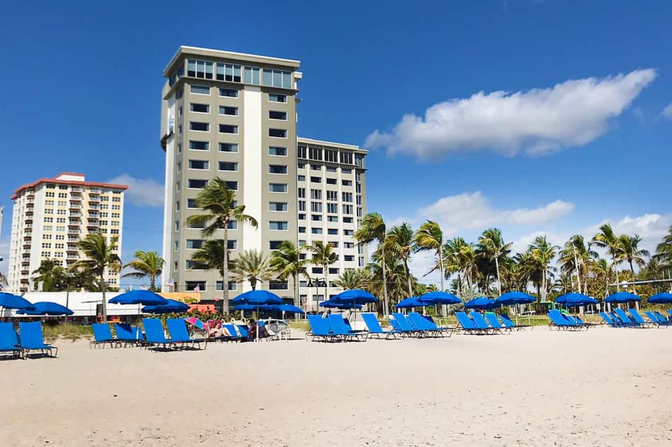 Beachfront hotel tower with blue umbrellas and lounge chairs on sandy beach, palm trees, and clear sky