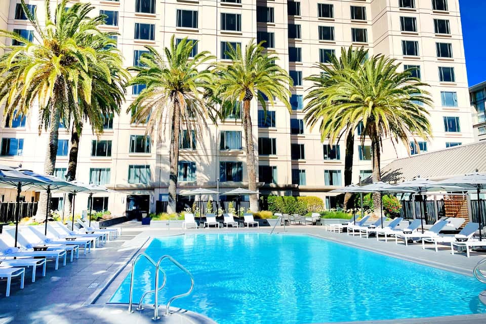 Resort pool with white loungers, palm trees, high-rise hotel building, and shade umbrellas