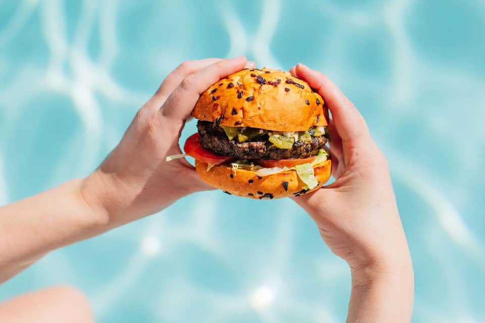 Hands holding gourmet burger with sesame seed bun over swimming pool water