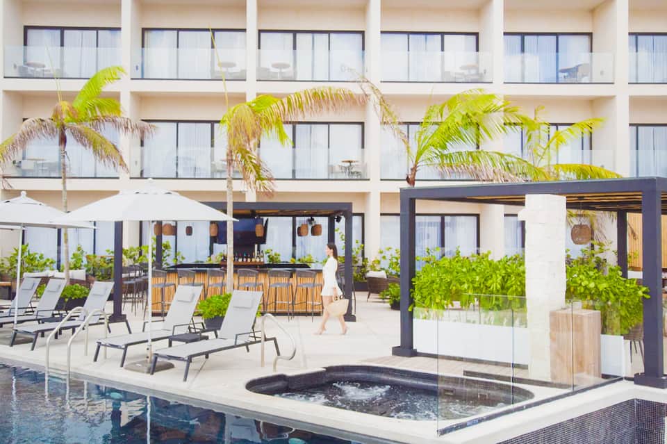 Modern resort courtyard with lounge chairs, pergolas, bar area, hot tub, and multi-story hotel building