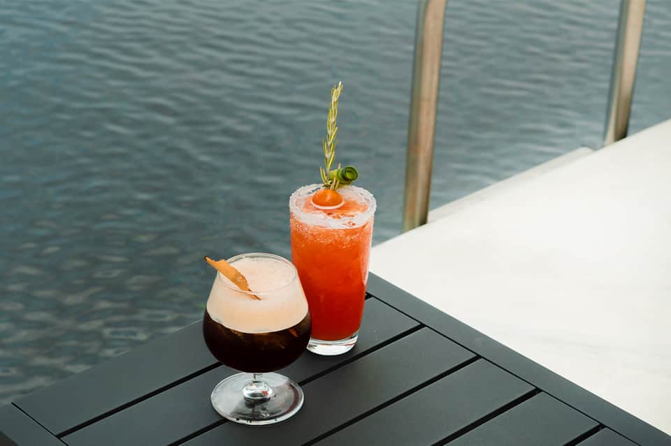 Two colorful cocktails on dark table overlooking calm waterfront at sunset