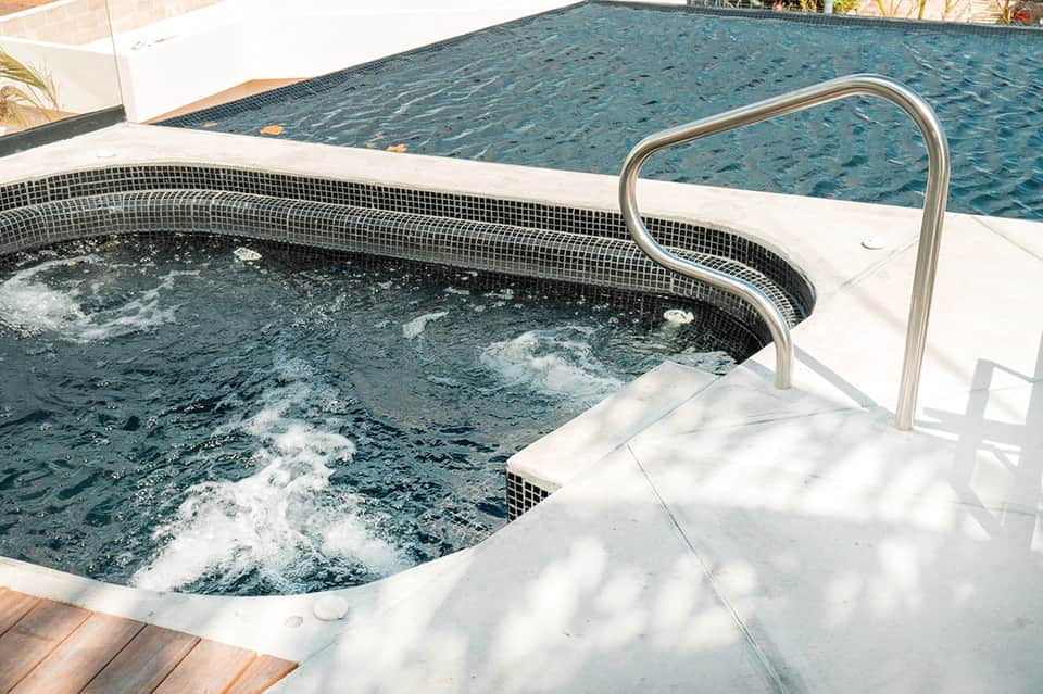 Outdoor spa hot tub with black mosaic tiles next to a larger swimming pool