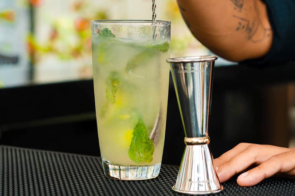 Bartender pouring mojito cocktail with mint and lime into glass on dark bar counter