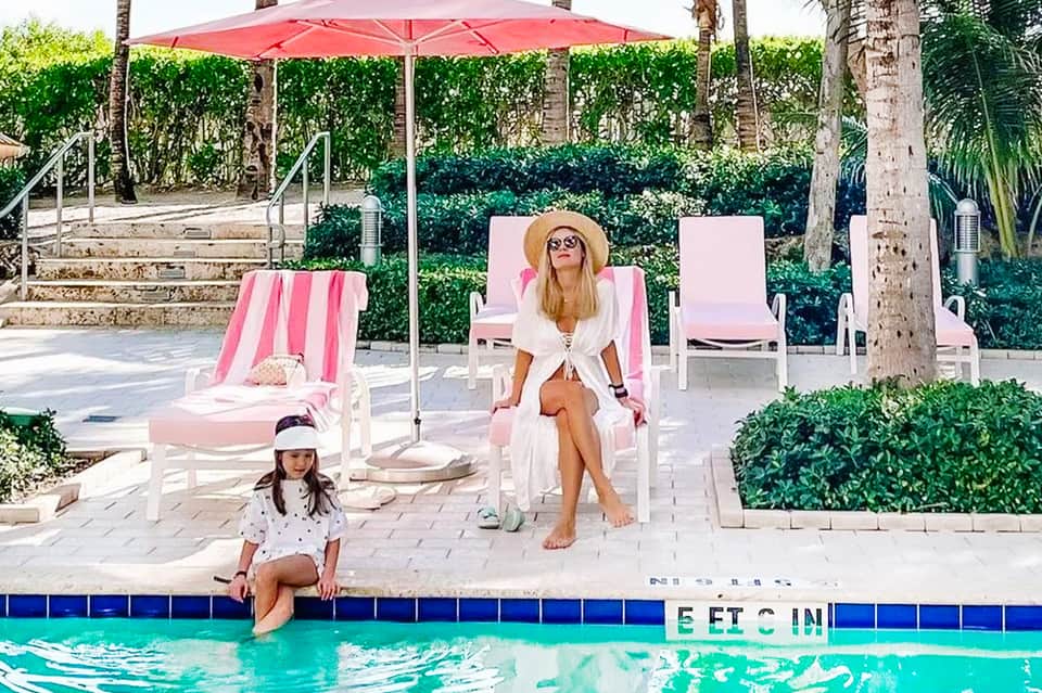 Woman and child relaxing poolside with pink lounge chairs and red umbrella in tropical setting