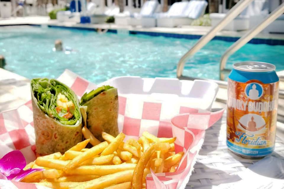 Poolside lunch setup with wrapped sandwich, french fries, and beverage on checkered paper