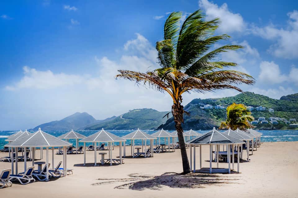 Beachfront cabanas with white umbrellas and lounge chairs on sandy beach with mountains in the background