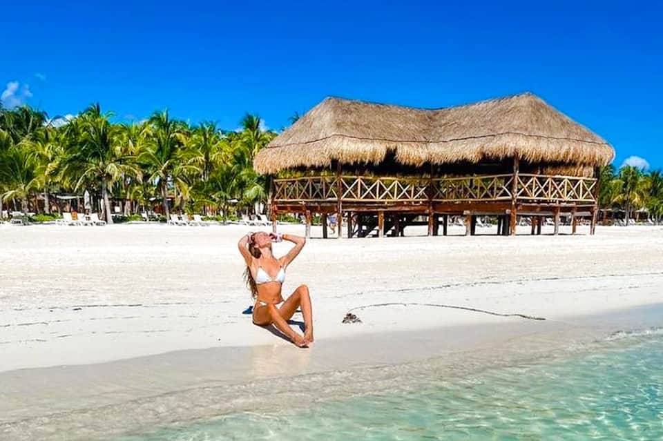 Woman relaxing on pristine white sand beach with thatched-roof beach club and palm trees in tropical setting