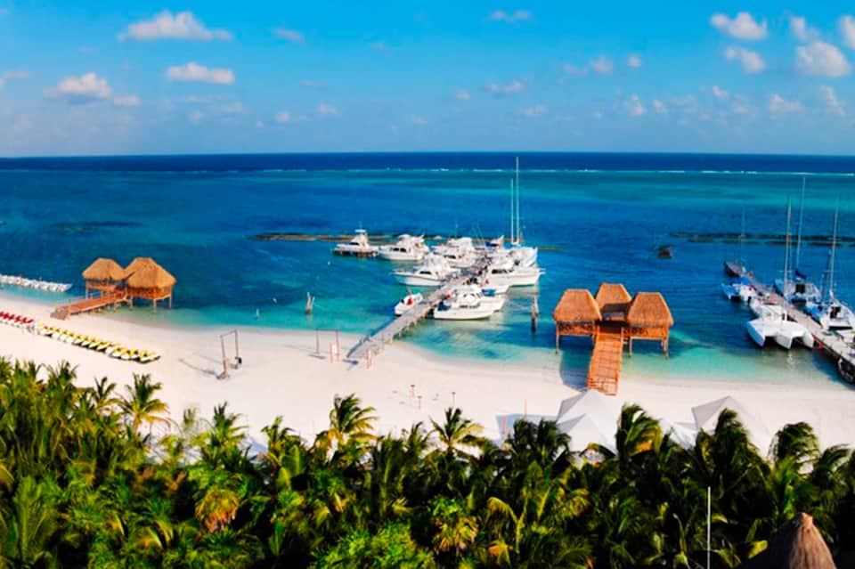 Aerial beach resort view with thatched bungalows, white sand, turquoise water, sailboats, palms