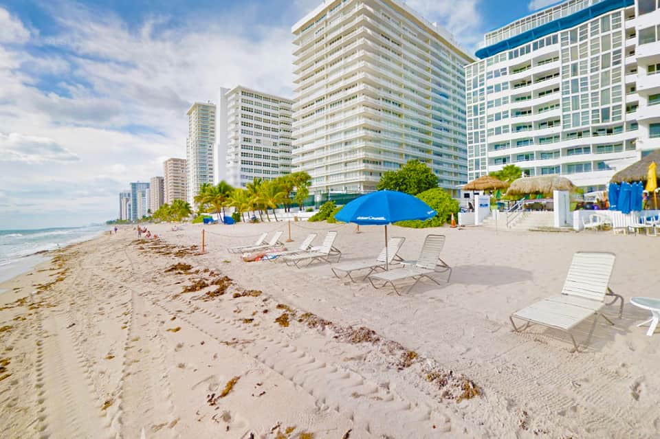 Beach scene with loungers and blue umbrellas, high-rise hotels, palm trees, and ocean