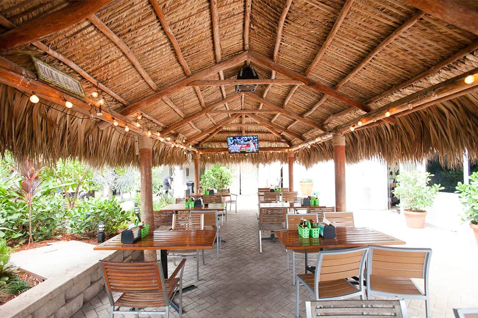Covered outdoor dining pavilion with thatched roof, wooden tables, chairs, and tropical landscaping