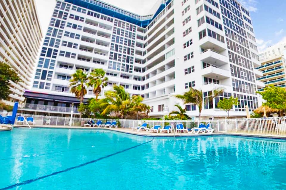 Resort swimming pool surrounded by high-rise residential buildings with palm trees and lounge chairs