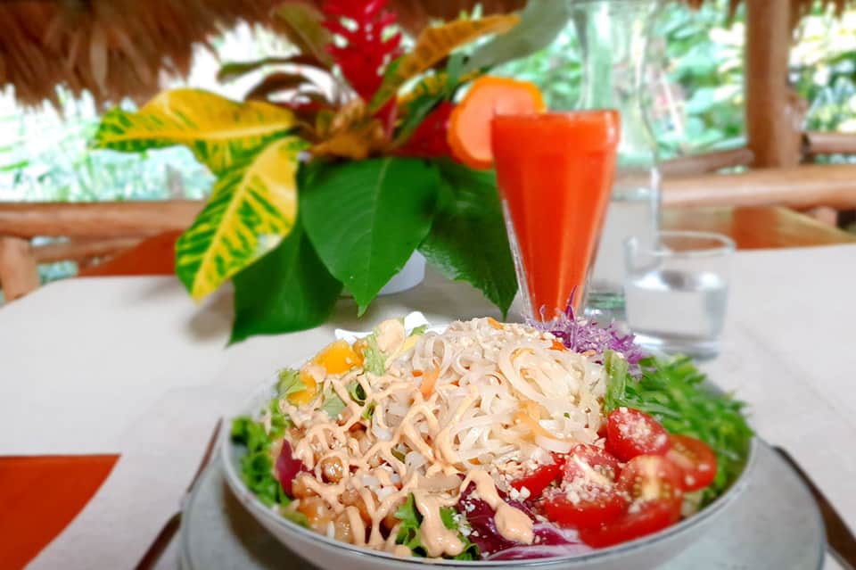 Beachside resort restaurant with noodle dish, tropical flowers, and thatched roof structures