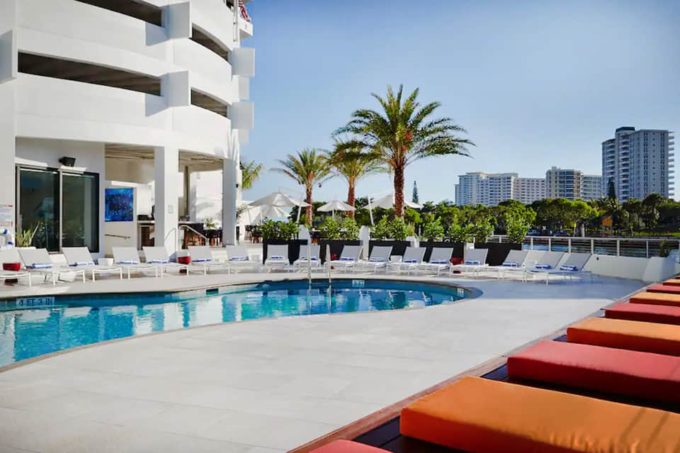Contemporary pool deck with colorful lounge cushions, palm trees, white building, and highrise hotels
