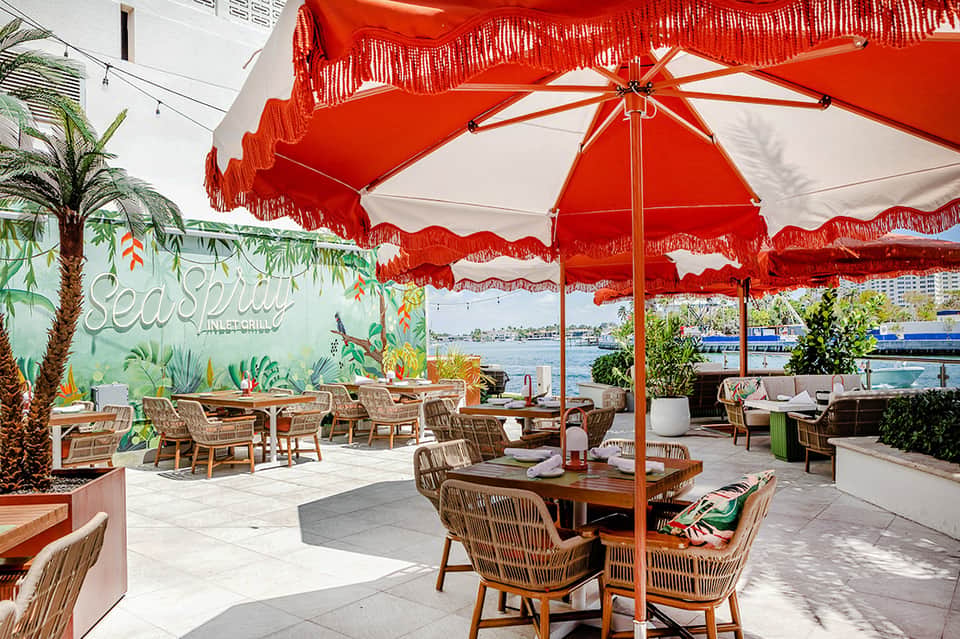 Waterfront restaurant patio with red umbrellas, woven chairs, tropical plants, and city skyline views