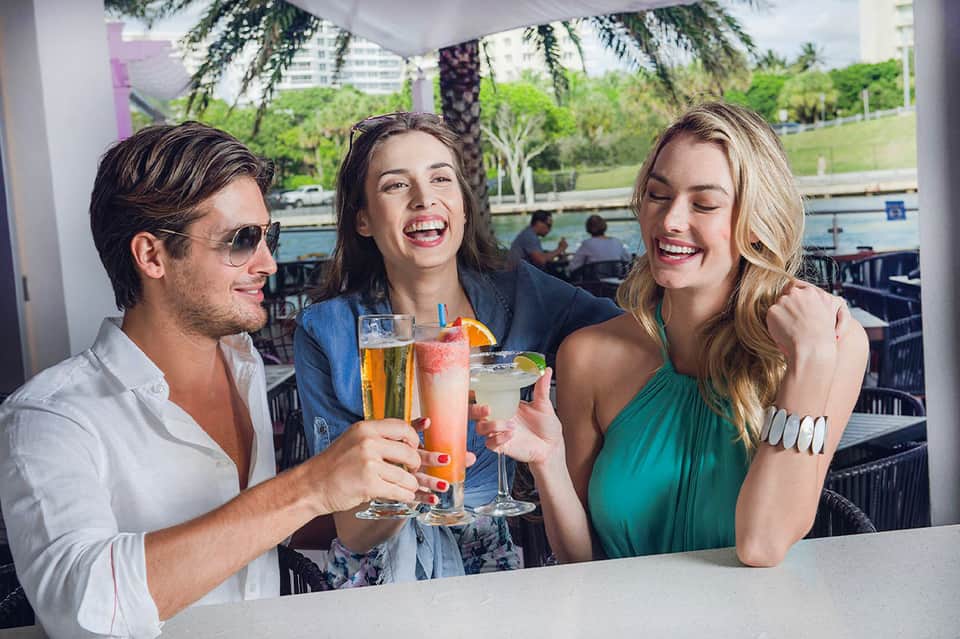 Three people toasting with cocktails at a waterfront bar overlooking a pool and palm trees