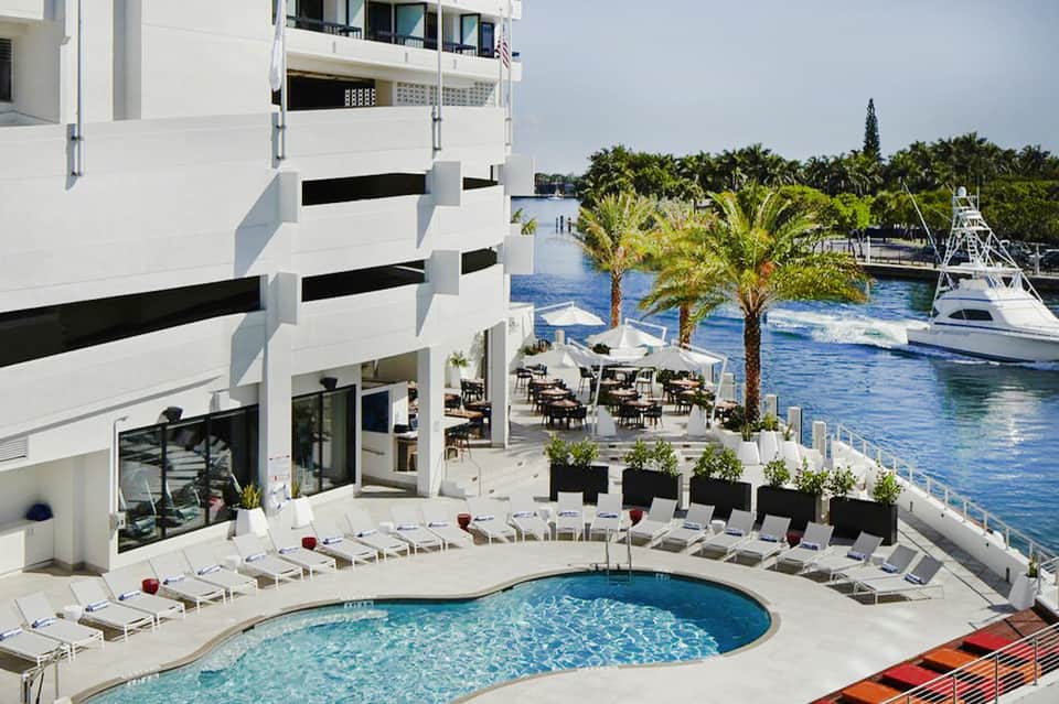 Waterfront pool deck with spiral building, palm trees, lounge chairs, and boats docked in blue-green water