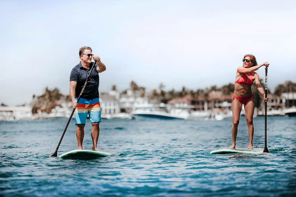 Two people stand-up paddleboarding on calm water with coastal residential area behind them