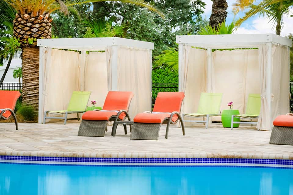 Resort pool deck with orange and lime lounge chairs under white cabanas surrounded by palms