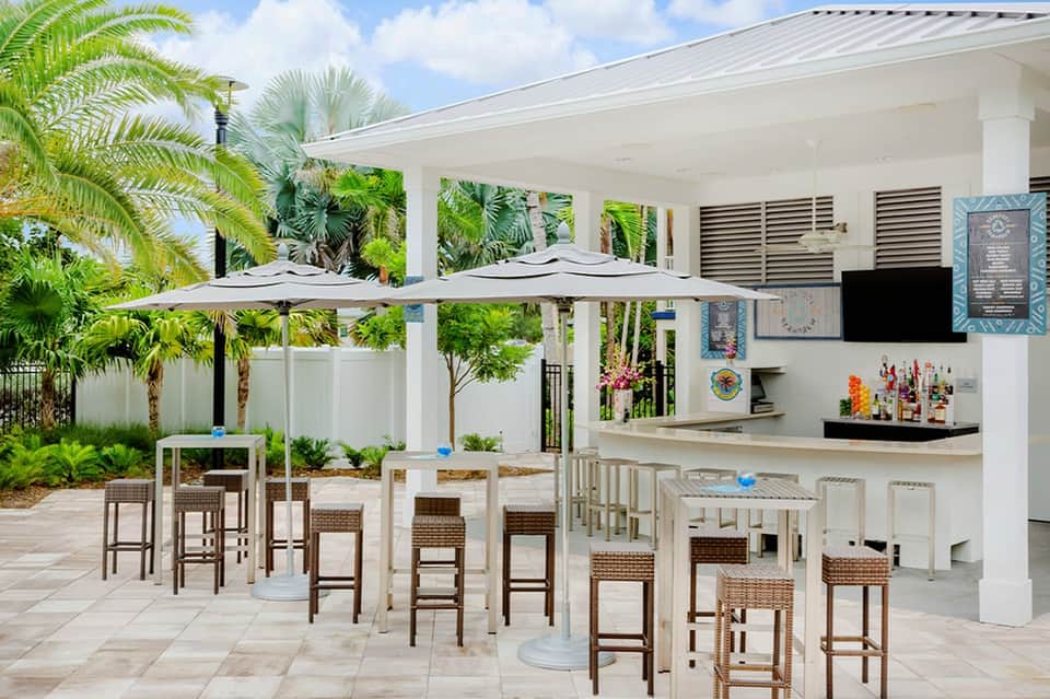 Outdoor poolside bar and kitchen area with white pergola, palm trees, and high-top seating